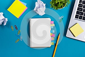 Office table desk with set of colorful supplies, white blank note pad, cup, pen, pc, crumpled paper, flower on blue