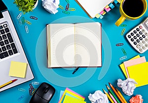 Office table desk with set of colorful supplies, white blank note pad, cup, pen, pc, crumpled paper, flower on blue