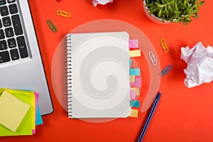 Office table desk with set of colorful supplies, white blank note pad, cup, pen, pc, crumpled paper, flower on red