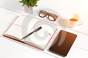 Office table desk with set of supplies, white blank notepad, cup, pen, tablet, glasses, flower on white background. Top