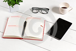 Office table desk with set of supplies, white blank notepad, cup, pen, tablet, glasses, flower on white background. Top