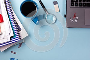 Office table desk with supplies, blank note pad, cup, pen, pc, crumpled paper, flower on blue background. Top view