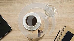 Office table with notepad, laptop, smart phone, coffee cup, glass of water and dark chocolate. Top view.