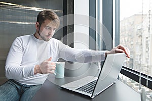 An office worker with a cup of hot drink in his hands begins to work on a laptop.