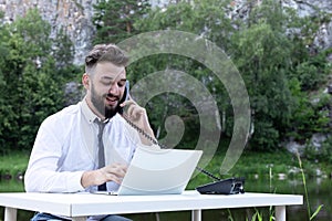 Office worker guy at desk using desktop computer laptop and phone at work, smiling, looking at screen. Outdoor