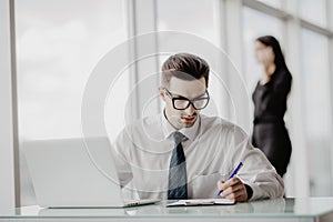 Office worker sitting at desk in office using laptop, while women talking phone in the background.