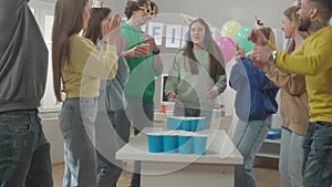 Office workers in holiday hats have fun playing beer pong. A young woman takes aim, throws a ball and misses, drinks