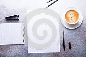 Office workplace with blank paper, cup of coffee, note pad and pen. Stone background. Top view