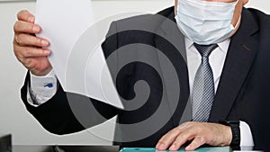 An official or businessman in a suit, tie and medical mask sits at a table and holds an important document in his hands