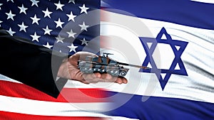 Official hand holding toy tank against USA and Israel flag, defense deal