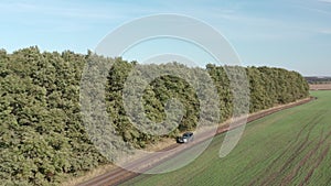 Offroad car driving along green trees on soil route and agricultural field in countryside. Drone view SUV auto moving on