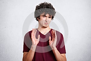 Oh no, don`t bother me! Annoyed grumpy male with crisp hair, shows refusal gesture, poses against white background. Curly handsome