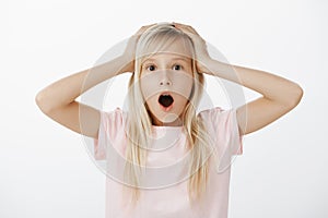 Oh no, I am in trouble. Studio shot of confused nervous young adorable girl with blond hair, dropping jaw and gasping