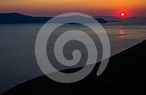 Oia santorini island greece. Natural coloured sunset. Dark red sky. Gold setting sun behind mountains. Calm sea reflecting