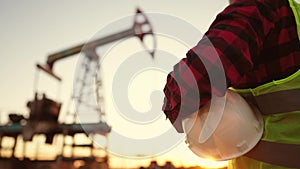 oil production. a worker holding a protective hard hat at sunset in the background an oil pump. oilfield business a