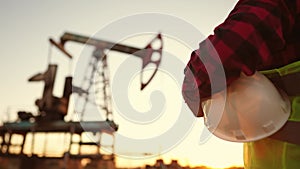 oil production. a worker holding a protective hard hat at sunset in the background an oil pump. oilfield business a