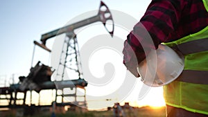 oil production. a worker holding a protective hard hat at sunset in the background an oil pump. oilfield business a