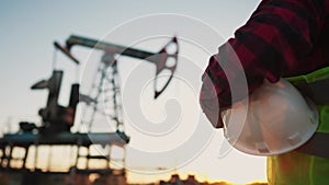 oil production. a worker holding a protective hard hat at sunset in the background an oil pump. oilfield business a