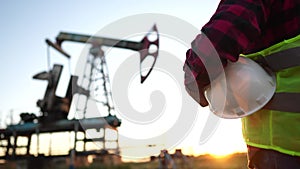 oil production. a worker holding a protective hard hat at sunset in the background an oil pump. oilfield business a