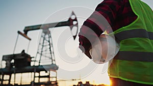 oil production. a worker holding a protective hard hat at sunset in the background an oil pump. oilfield business a