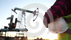 oil production. a worker holding a protective hard hat at sunset in the background an oil pump. oilfield business a