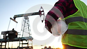 oil production. a worker holding a protective hard hat at sunset in the background an oil pump. oilfield business