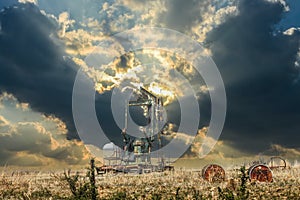 Oil well pump jack in field with old oil barrels behind barbed wire fence under dramatic sunset sky with dark oninous clouds