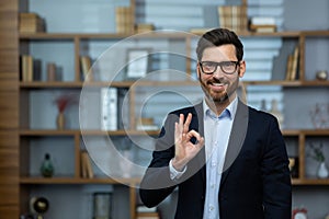 OK sign, businessman shows approval sign to camera, smiling man in business suit inside office at workplace, experienced