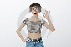 Okay, I agree. Portrait of charming stylish urban girl in striped cropped top and round glasses, raising hand on ok