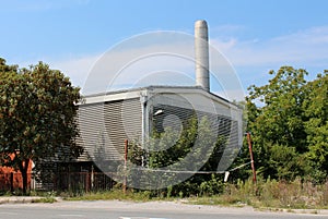 Old abandoned factory with red brick wall and metal insulation surrounded with large trees and overgrown vegetation