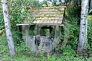 Old abandoned forest well with wooden frame and picket fence covered with red roof tiles surrounded with dense vegetation and