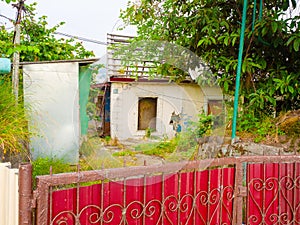 An old, abandoned and overgrown with grass one-story house with a destroyed roof and broken windows behind a fence made