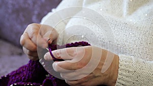 Old adult woman is sitting on the sofa and knitting. Close-up of women`s hands doing handicraft handwork, hobby of adult women in