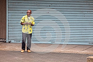 Old African man standing against grey security shutters looking to camera, full length