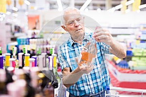 Old age man examines bottle of vermouth in alcoholic section of supermarket