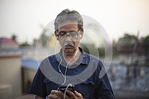 An old / aged thoughtful Indian Bengali man in blue shirt is listening to music in his mobile phone using white earplugs