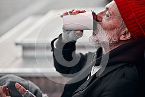 Old-aged vagabond sitting on walking street and drinking coffee or tea