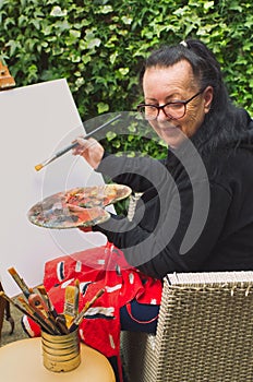 Old artist lady holding painter pallete, smiling and starting to paint on the empty canvas