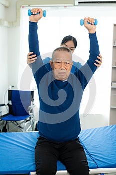 An old Asian man is doing physical therapy with the support of a nurse in the living room by exercising