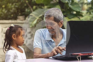 An old asian man in his 70s listens to his curious granddaughter while typing on a laptop while sitting outside at his garden