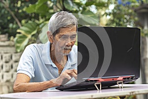 An old asian man in his 70s typing on a laptop while sitting outside at his garden