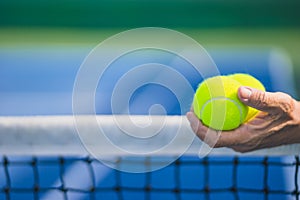 Old asian man hold two tennis balls in left hand, selective focus, blurred net and blue and green tennis court as backgroun