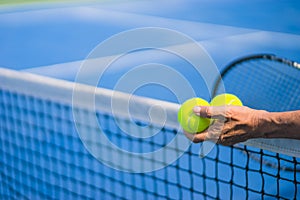 Old asian man hold two tennis balls in left hand, selective focus, blurred racket, net and green tennis court as background