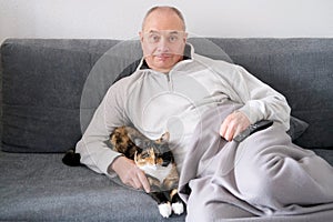 Old bald man sitting on the sofa under a blanket with a cat, TV remote control, concept of cozy home evening, family life