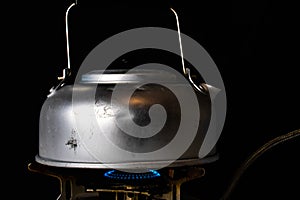 Old banged up steel kettle boiling water on a camping gas stove with a blue flame. Isolated on black background. photo