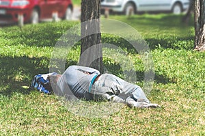 Old barefoot homeless or refugee man sleeping on the grass in the city park using his travel bag as pillow, social documentary str