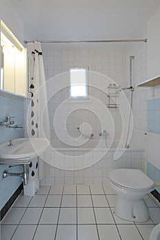 Old bathroom with white tiles, at the end there is a bathtub with shower and open curtain. There is also a small window