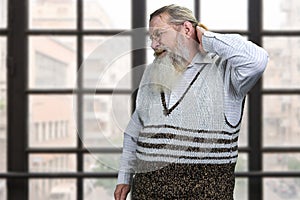 Old bearded man suffering from nape pain standing on windows background.