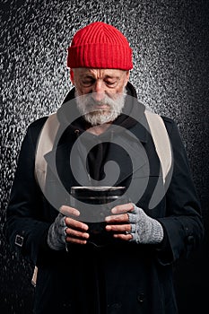 Old beggar man in street clothes stand under rain look with sadness at iron cup