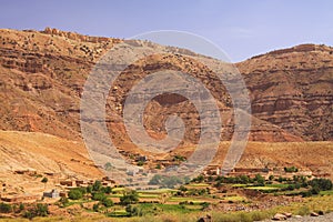 Old berber village oasis with houses build of clay bricks in front of impressive high rugged red mountain face, Gorges du Dades,